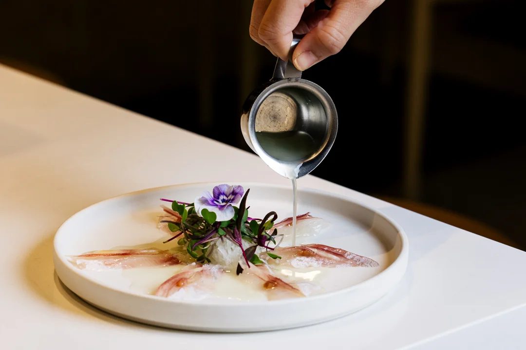 Chef pouring sauce over a plate of sashimi garnished with microgreens and a viola flower at Harumi Sushi & Sake